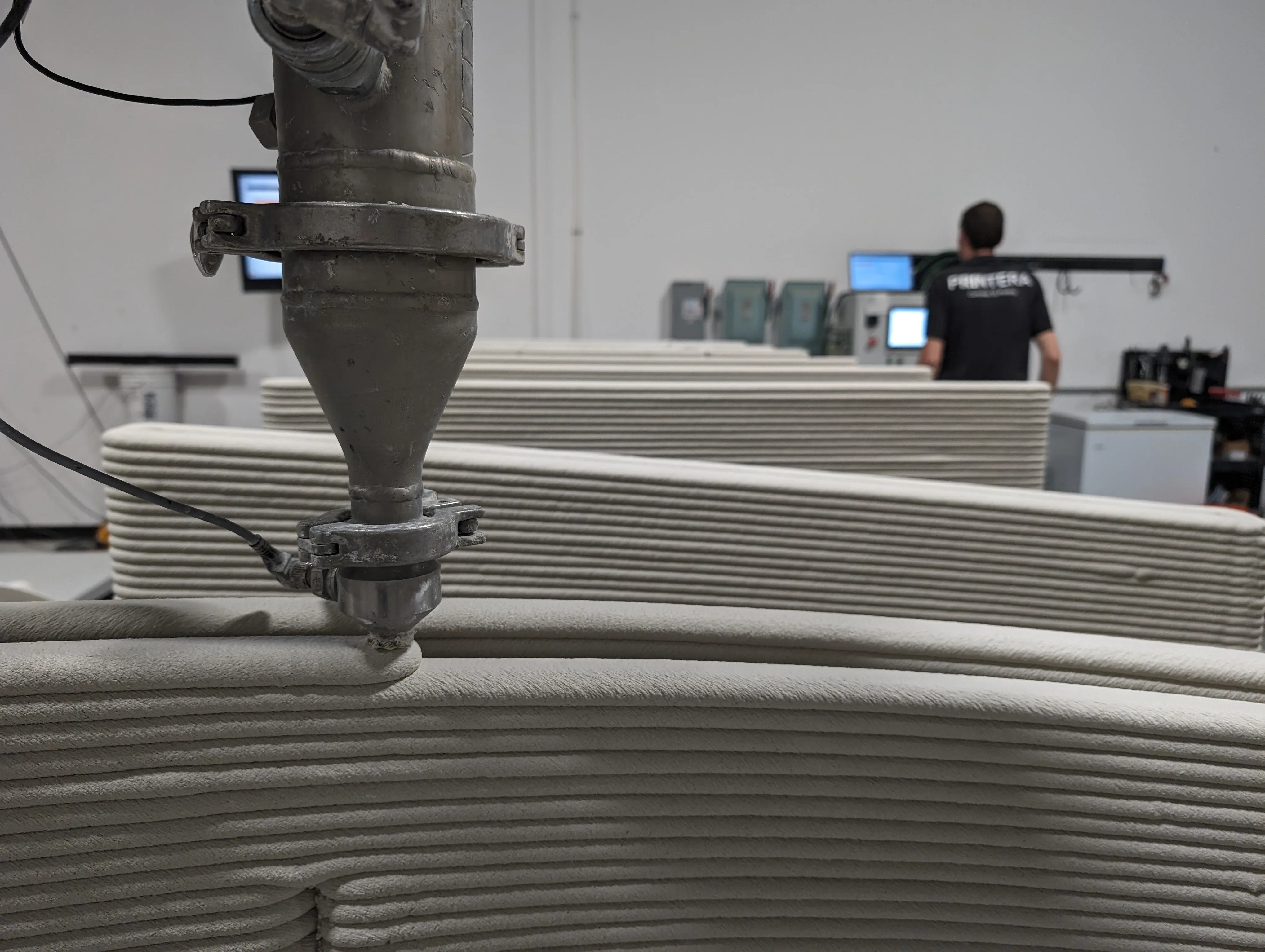 Robotic arm using 3D concrete printing to produce small wall segments in a controlled facility environment.